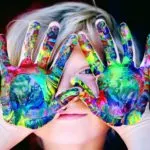 A child's face peeking through colorful paint-covered hands